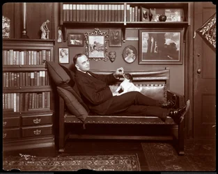 Will R. Barnes reclined on a couch with a dog in his library studio, 1903
